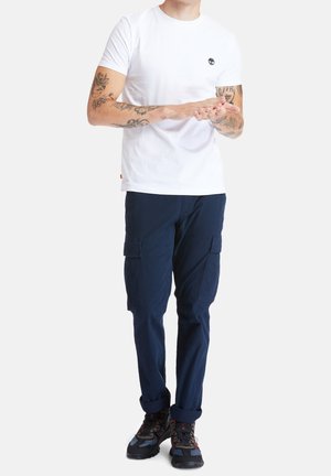 White cotton t-shirt with a small logo on the chest, paired with navy cargo pants featuring side pockets and rolled cuffs. Black hiking shoes.