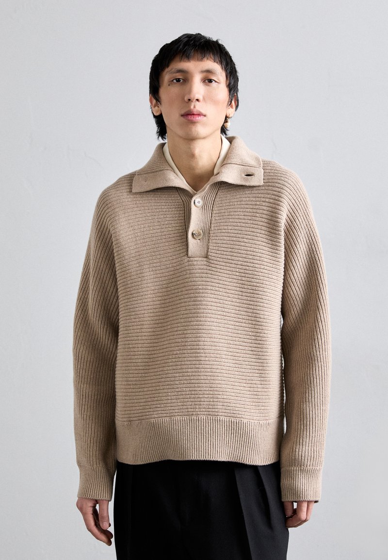 Beige, ribbed knit pullover with a collar, featuring three buttons and a textured finish. Long sleeves and a relaxed fit.