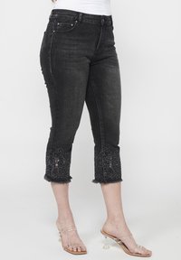 Crne traperice do članka s resama, sa vezenim detaljima na manžetama. Klasičan dizajn s pet džepova i prilagođenim siluetom.