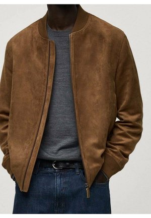 Homme portant une veste bomber en daim marron sur un pull gris, les mains dans les poches et un jean bleu foncé avec une ceinture noire.