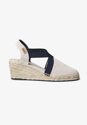 Beige vászon espadrille, tengerészkék elasztikus keresztpántokkal és fonott juta sarokkal. Gumi talppal rendelkezik, amely biztosítja a tapadást és a kényelmet.