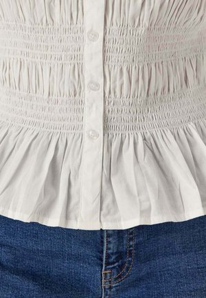 Witte geplooide blouse met knopen gedragen met blauwe spijkerbroek, met gedetailleerde textuur en stofplooien.