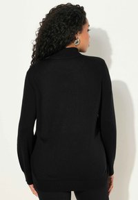 Zwarte trui gemaakt van een zacht, gebreid materiaal. Heeft een hoge kraag en lange mouwen, met een iets losse pasvorm en zijsplitten aan de zoom.