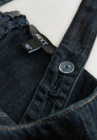 Mörka jeansfoder med dragkedjestängning, plastknapp och en svart etikett med "NEXT," mot en ljus bakgrund.