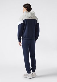 Uomo in piedi di spalle, che indossa un felpa navy e grigia con strisce bianche e pantaloni jogger navy abbinati, con sneaker bianche.