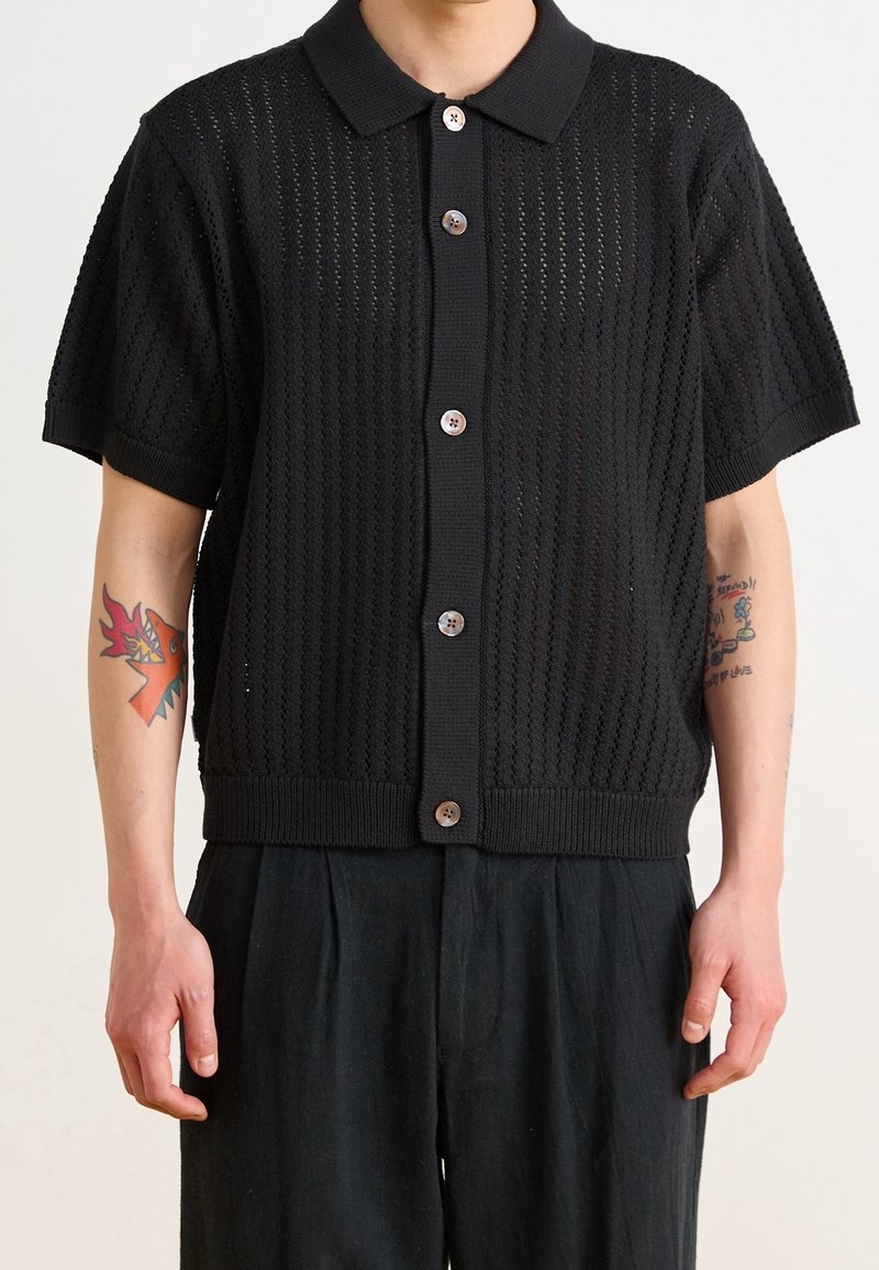 Černý krátký pletený kardigan s krátkými rukávy a texturovaným vzorem, zapínáním na knoflíky vpředu a límečkem. Má dřevěné knoflíky a žebrovaný lem.