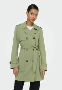 Femme portant un trench-coat vert clair à double boutonnage avec une ceinture nouée à la taille, debout devant un fond clair uni.