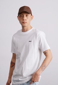 Hvit bomulls T-skjorte med "OBEY" logo i svart, kombinert med en brun caps. Modellen har synlige tatoveringer på armene. Enkelt, uformelt design.