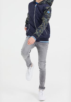 Kind trägt eine marineblaue gesteppte Jacke mit Camouflage-Ärmeln und Kapuze, hellgraue Jeans und weiße Turnschuhe, hebt beim Gehen ein Bein nach vorne.