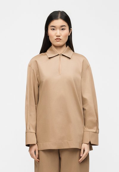 Femme aux longs cheveux noirs portant une chemise ample beige à manches longues avec un col à boutons et un pantalon beige assorti, debout devant un fond blanc.