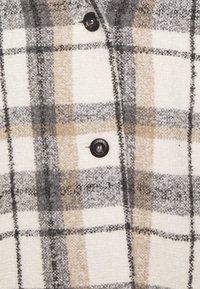 Camicia a quadri in tessuto intrecciato crema, grigio e beige. Presenta bottoni neri e una superficie testurizzata con fibre e cuciture visibili.