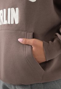 Mano con smalto rosa nella tasca di una felpa marrone con la scritta bianca ricamata "BERLINO", che mostra il polsino della manica e parte dei pantaloni grigi.