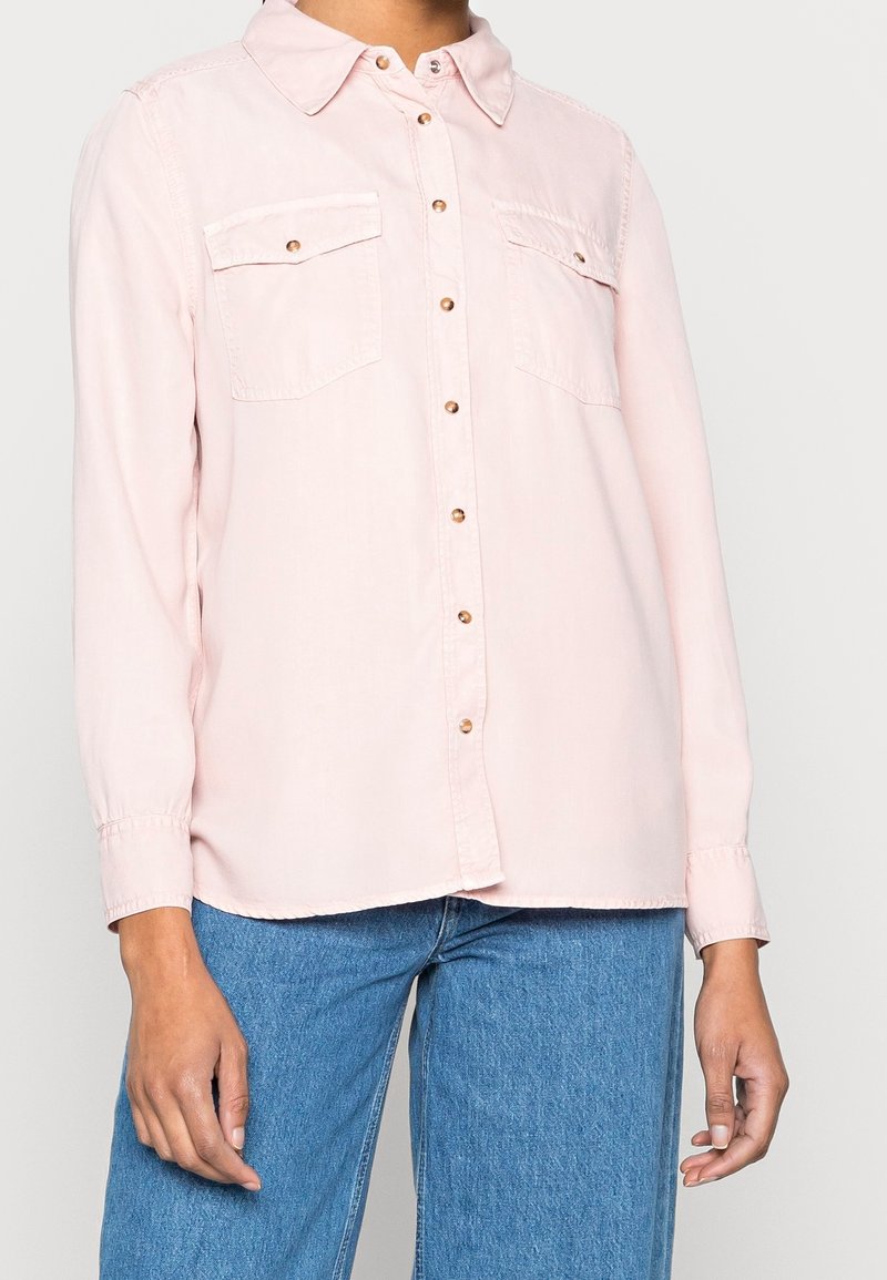Chemise rose clair à manches longues avec fermeture boutonnée sur le devant, deux poches poitrine et boutons dorés ; dotée d'un col à revers et d'une coupe décontractée.
