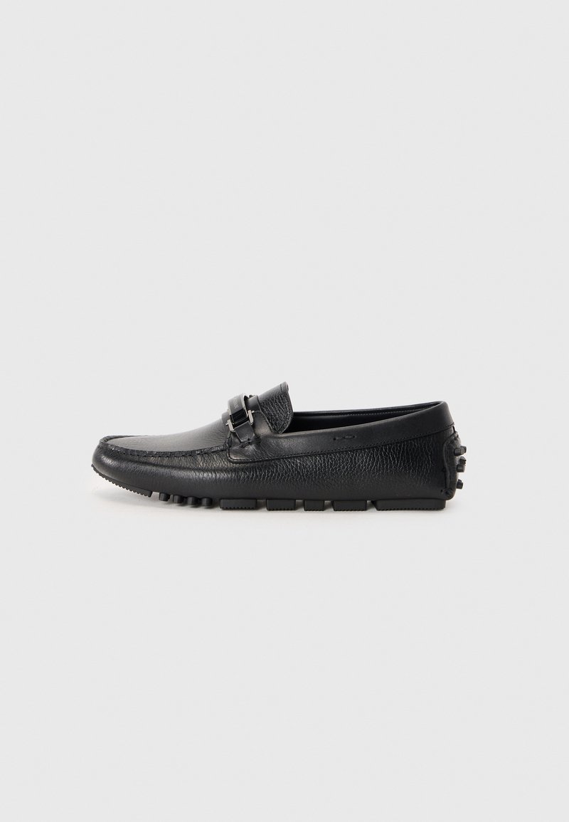 Mocasines de cuero negro con acabado texturizado, una hebilla decorativa y suela de goma con acentos salientes. Vista lateral mostrada.