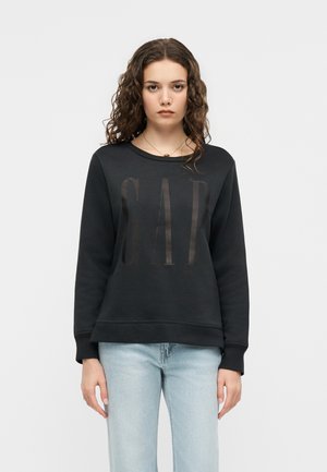 Tânără cu păr creț poartă un hanorac negru GAP și blugi bleu deschis, stând pe un fundal alb simplu.