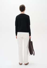 Person som står med ryggen mot kameran, iklädd svart tröja, vita byxor, beige skor, hållande en mörkbrun tygväska i höger hand mot en vit bakgrund.