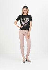 Crna pamučna majica s grafikom "Texas" i dizajnom orla, u kombinaciji s svijetloružičastim uskim trapericama i bež visokim potpeticama.