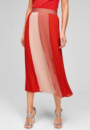 Vrouw draagt een geplooide midirok met verticale panelen in rood, roze en koraal, gecombineerd met zilverkleurige sandalen met hoge hakken.