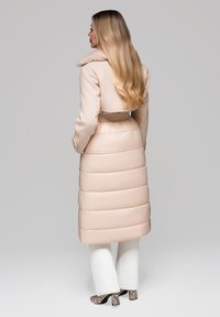 Žena s dlouhými blond vlasy, oblečená v béžovém prošívaném kabátu s kožešinovým límcem, bílých kalhotách a lodičkách s hadím vzorem, stojící bokem.