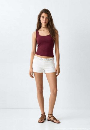 Ung kvinde iført en ærmeløs burgunder top, hvide shorts med nitter og brune sandaler med nitter står mod en ensfarvet hvid baggrund.