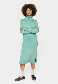 Tirkīza adīta turtleneck kleita ar garām piedurknēm, sānu šķirtni un ribotām aprocēm. Saskaņota ar pelēkām zeķēm un brūnām loafers.