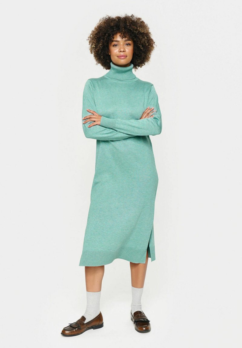 Tirkīza adīta turtleneck kleita ar garām piedurknēm, sānu šķirtni un ribotām aprocēm. Saskaņota ar pelēkām zeķēm un brūnām loafers.