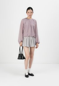 Woman wearing a sheer patterned long-sleeve blouse, pleated gray mini skirt, white socks, black flats, holding a black handbag.