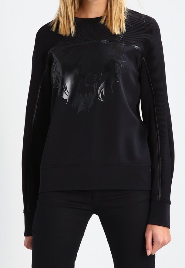 Femme portant un top noir à manches longues avec des panneaux texturés et un motif de lion en relief sur la poitrine, associé à un pantalon noir.