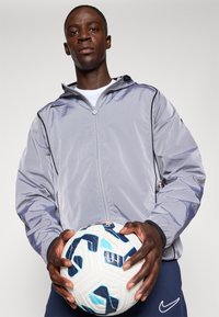 Grå lättviktsjacka med dragkedja fram och huva, med svarta detaljer. Individen håller en vit och blå mönstrad fotboll.