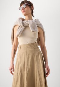 Débardeur beige associé à une jupe kaki plissée taille haute. Pull léger en tricot beige clair drapé sur les épaules, avec des lunettes de soleil.