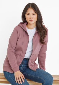 Pink zip-up hoodie with a hood, side pockets, and drawstrings, made of soft fabric, paired with a white t-shirt and dark jeans.