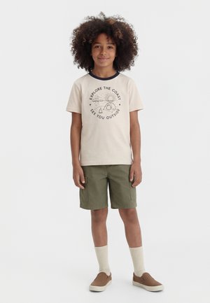 Copil cu păr creț purtând un tricou bej cu grafică de camping la malul mării, pantaloni scurți oliv, șosete înalte crem și pantofi slip-on maro, stând pe un fundal alb.