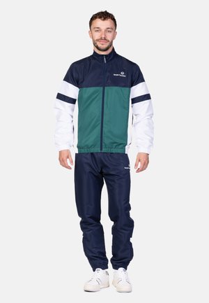 Farbblockierter Jogginganzug bestehend aus einer dunkelblauen, grünen und weißen Jacke mit Reißverschluss, kombiniert mit dunkelblauen Hosen und weißen Sneakers.