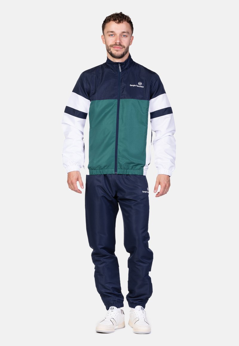 Tuta a blocchi di colore composta da una giacca blu navy, verde e bianca con chiusura zip, abbinata a pantaloni blu navy e sneakers bianche.
