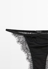 Tanga in pizzo nero con motivo floreale, dotato di bordi in pizzo trasparente e corpo in tessuto liscio. Etichetta che indica "Massimo Dutti."
