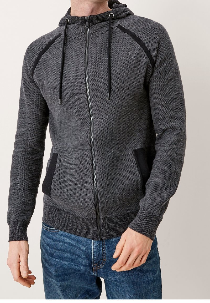 Homme portant un sweat à capuche gris foncé avec une fermeture éclair, cordons et accents noirs, associé à un jean en denim bleu, sur un fond uni.