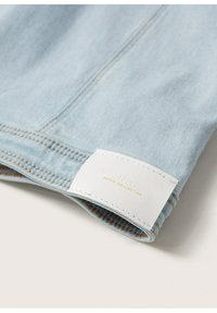 Ljust blå denimtyg med synlig sömnad. Inkluderar en vit etikett med "MNG DENIM COLLECTION" i guldtext.