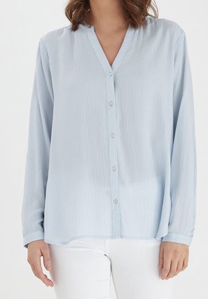 Vrouw die een lichtblauw, verticaal gestreept blouse met knopen en lange mouwen en een witte broek draagt, zichtbaar vanaf midden borst tot heupen.