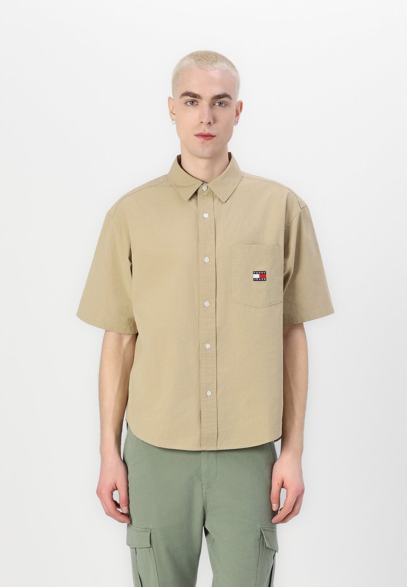 Chemise beige à manches courtes avec boutons, dotée d'une poche poitrine avec un petit logo et d'une coupe décontractée. Le tissu semble lisse et léger.