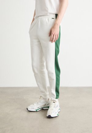 Mężczyzna w białych spodniach dresowych z zielonymi paskami po bokach, stojący w pomieszczeniu na szarej podłodze, z rękami w kieszeniach, w białych sneakersach.