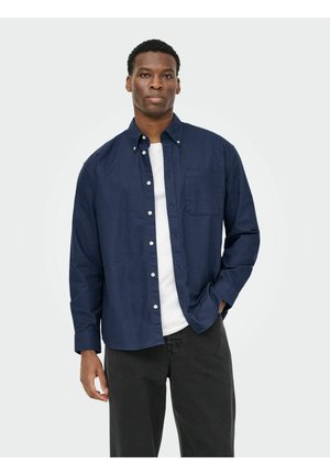 Bărbat stând, purtând o cămașă bleumarin cu nasturi peste un tricou alb și pantaloni negri, o mână în buzunar, pe un fundal simplu.
