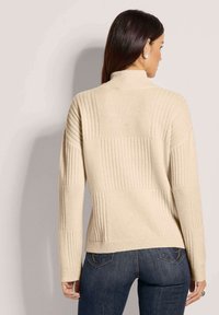 Beiger Pullover mit hohem Kragen und strukturierten Rippenmustern. Lange Ärmel und lockere Passform, kombiniert mit dunklen Jeans.