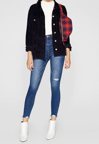 Marinblå kordjacka över en vit topp, matchad med slitna blå skinny jeans och vita ankelstövlar. Röd rutigt axelväska.