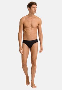 Zwarte, aansluitende zwemboxers voor mannen met een elastische tailleband, een gladde textuur en een minimalistisch ontwerp, gecombineerd met blote voeten.