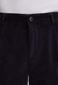 Marineblauwe corduroy broek met verticale ribbelstructuur, voor sluiting met knoop en klassieke snit met zakken. Minimalistisch ontwerp in neutrale kleuren.
