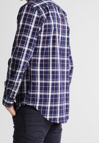 Niebiesko-biała koszula w kratę z czerwonymi akcentami, długimi rękawami i zaokrąglonym dołem. Wykonana z miękkiej tkaniny, z mankietami na guziki i kołnierzem.