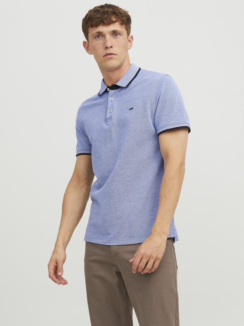 Blauwe polo met een drieknoopsluiting, zwarte kraag en contrasterende afwerking. De stof heeft een gestructureerde afwerking. Draagt met lichtbruine broek.