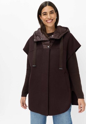 Femme souriante portant un manteau à capuche marron foncé sans manches superposé sur un haut à manches longues et un jean bleu sur fond blanc.