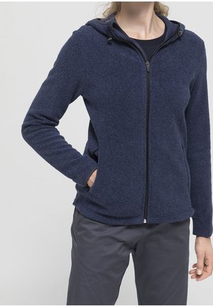 Osoba ubrana w granatową polarową kurtkę na suwak z kapturem i ciemne spodnie, jedna ręka w kieszeni, na jednolitym jasnym tle.