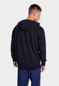 Zwarte hoodie van zachte stof met een trekkoordcapuchon, ribgebreide manchetten en zoom. Vanuit de achterkant getoond, gecombineerd met marineblauwe broek.